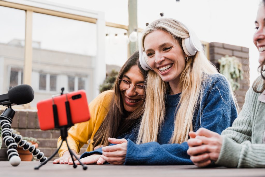 Happy friends streaming online podcast with smartphone outdoors in restaurant